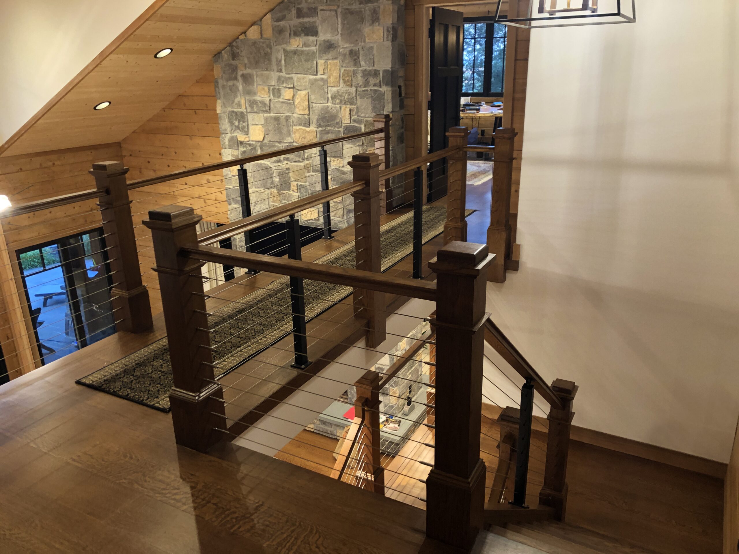 Rustic modern staircase with wood railings and cable wire design, overlooking stone accent wall and open-concept cabin interior.